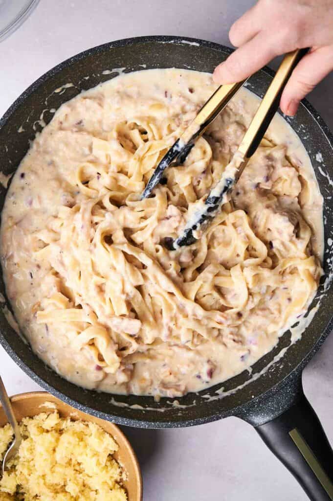 A skillet containing creamy tuna noodle casserole with a pair of tongs, accompanied by a side of grated cheese.