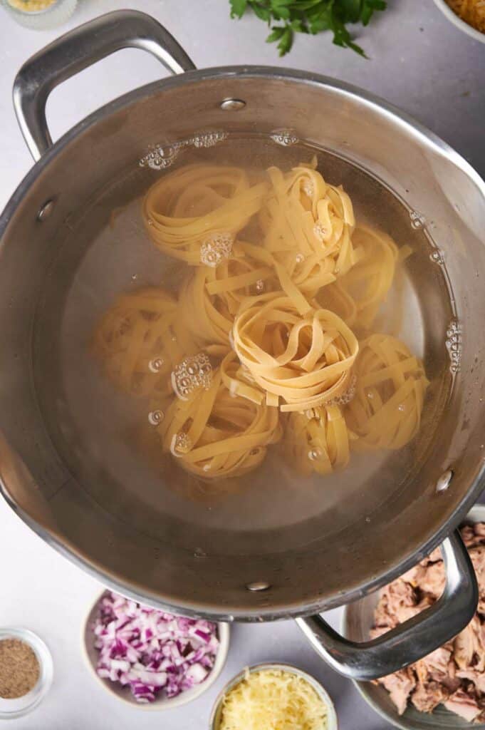 Cooked pasta in a pot of boiling water, viewed from above with ingredients like spices, chopped onions, and tuna around the countertop.