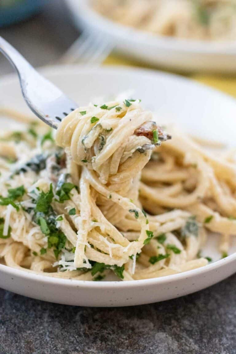 A fork is lifting a twirl of Creamy Tuscan Pasta garnished with chopped parsley and grated cheese from a white plate.