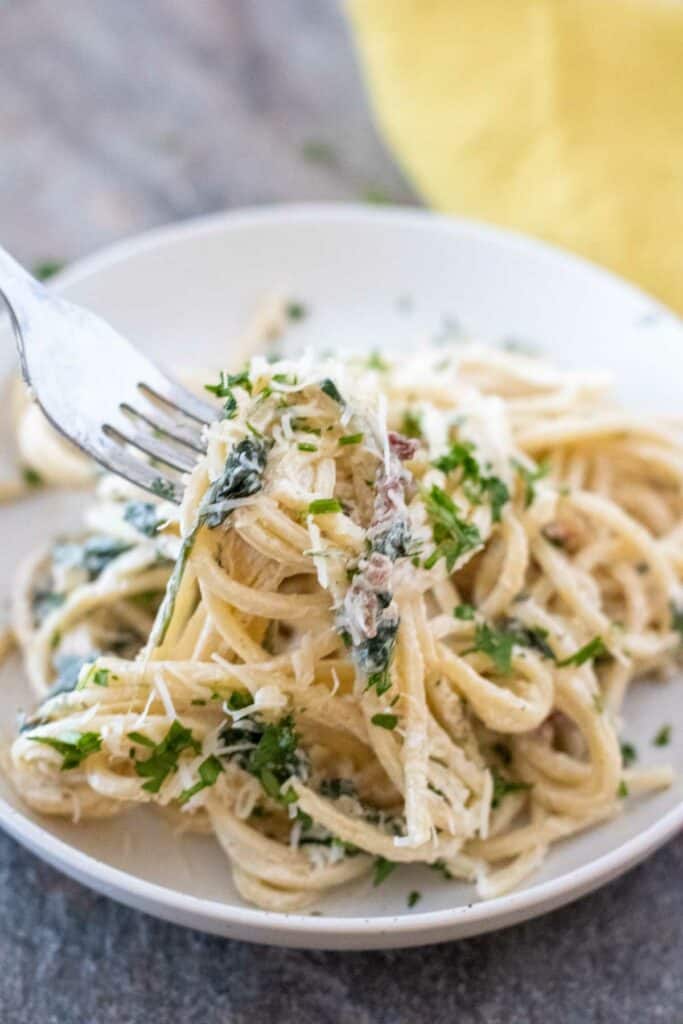 Fork twirling creamy Tuscan pasta with greens and grated cheese on a white plate, placed on a gray table with a yellow napkin in the background.