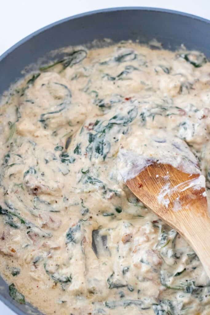 A creamy spinach and artichoke dip being stirred with a wooden spoon in a pan, looking just as indulgent as a Creamy Tuscan Pasta.