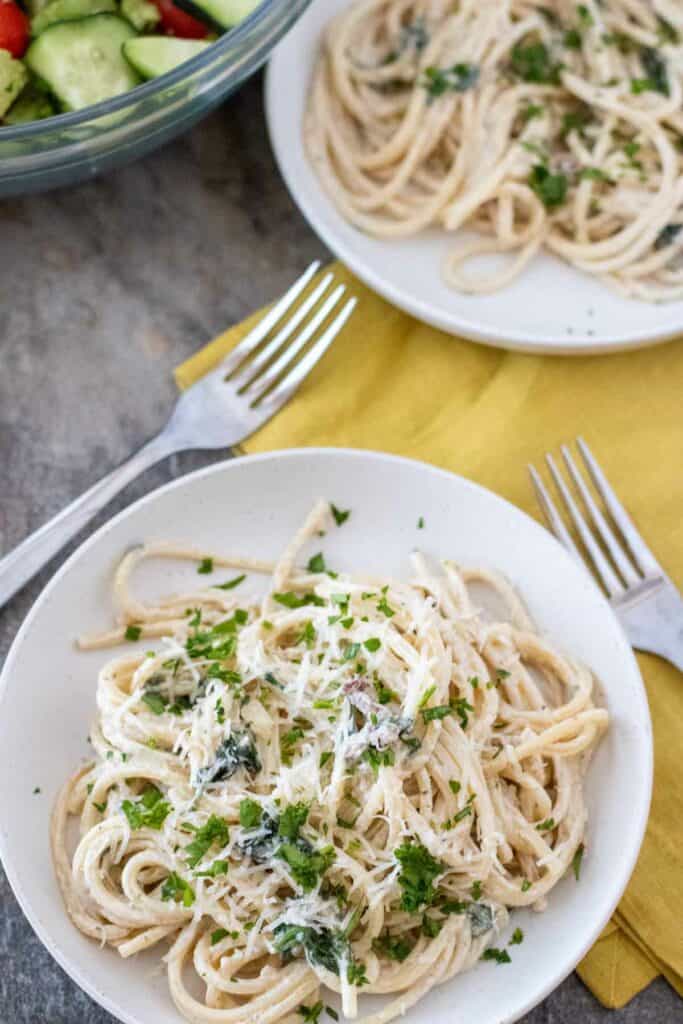 Two plates of creamy Tuscan pasta garnished with chopped herbs, with a side salad in the background. Two forks are placed on a yellow napkin next to one of the plates.