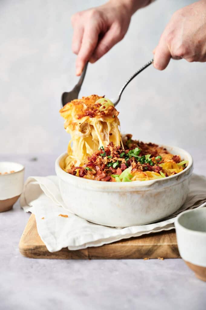 Hands serving an air fryer chicken casserole topped with bacon and green onions from a white baking dish on a wooden board.