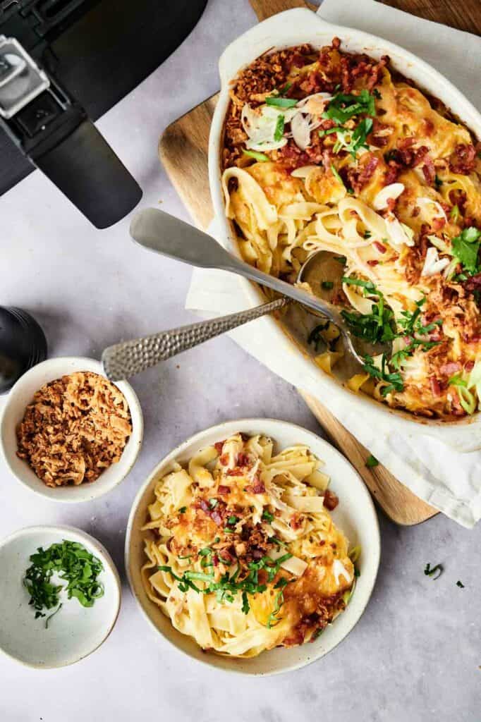 A top-down view of a table with two dishes of air fryer chicken casserole topped with bacon and herbs, garnished with a bowl of breadcrumbs and a small bowl of chopped herbs.