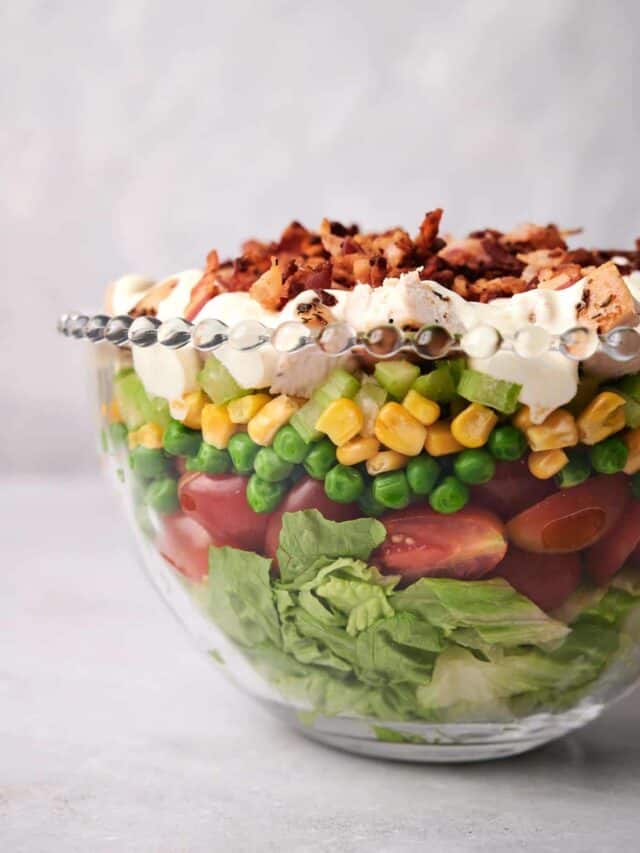 A layered salad in a clear bowl, featuring lettuce, cherry tomatoes, peas, corn, celery, cheese, and bacon bits, topped with a creamy dressing.