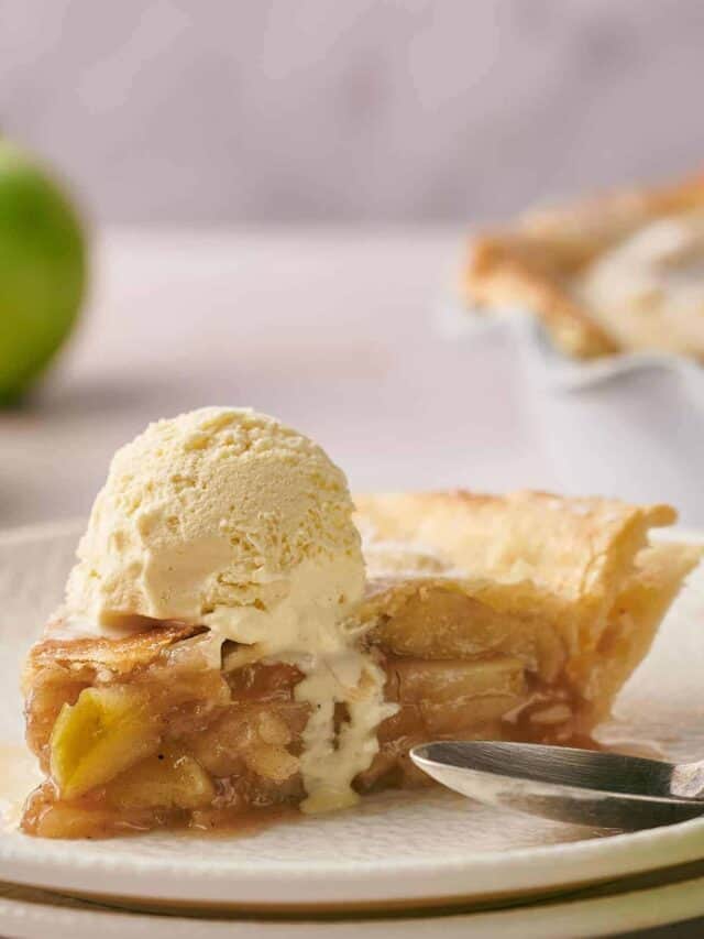 A slice of apple pie topped with a scoop of vanilla ice cream on a white plate with a stainless steel spoon resting beside it. A whole green apple is visible in the blurred background.