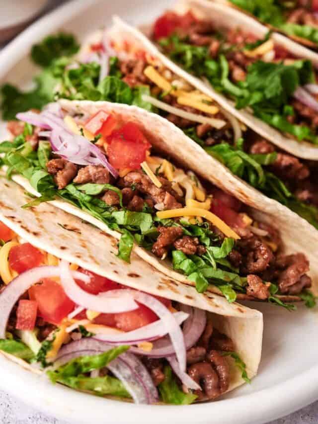 Three tacos filled with seasoned meat, shredded cheese, chopped tomatoes, sliced onions, and fresh chopped greens on a white plate.