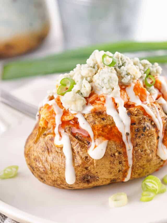 A baked potato topped with Buffalo sauce, blue cheese crumbles, and a drizzle of white dressing is on a white plate, garnished with sliced green onions.