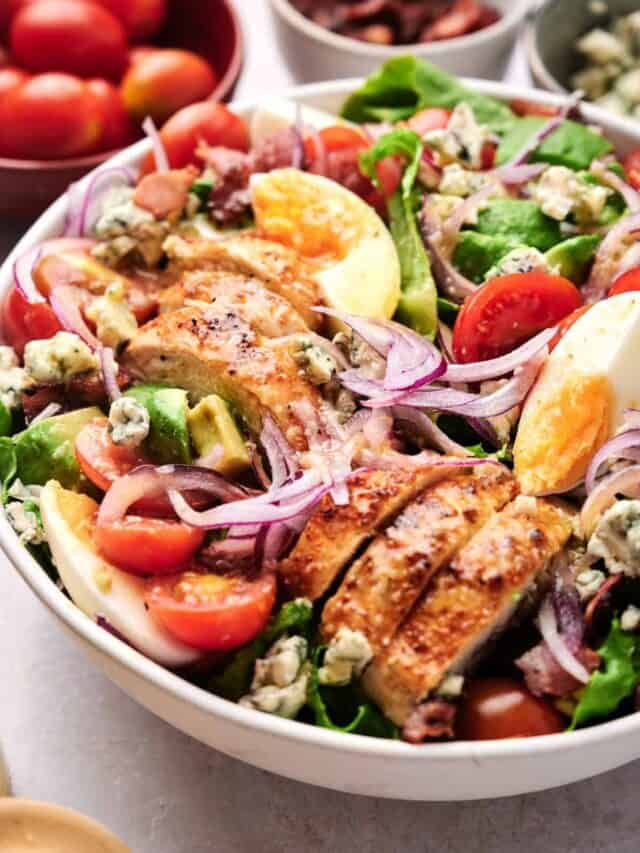 A bowl of salad with grilled chicken, avocado, tomatoes, red onions, gorgonzola cheese, hard-boiled eggs, and bacon bits on a bed of greens.