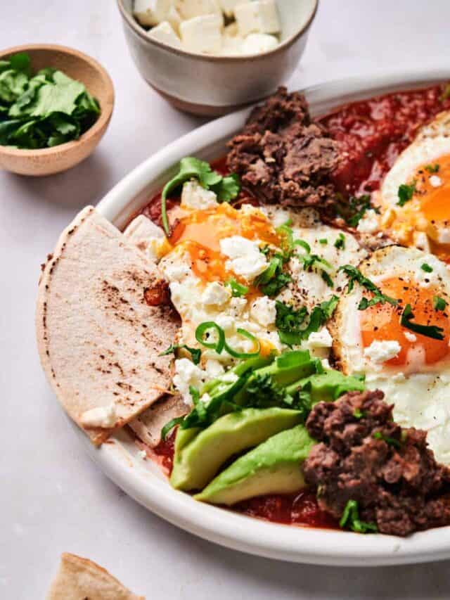 A plate with sunny-side-up eggs, avocado slices, refried beans, crumbled cheese, and green herbs. A bowl of cheese and a dish of greens are beside the plate. A piece of pita is on the plate's edge.