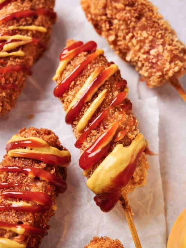 Close-up of crispy corndogs on sticks, drizzled with ketchup and mustard. They are placed on a parchment paper background.