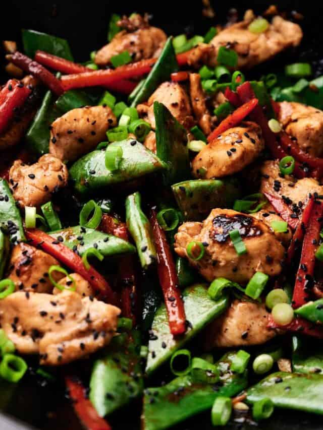 A close-up of a stir-fry dish with chicken, sugar snap peas, red bell peppers, and green onions, garnished with black sesame seeds.