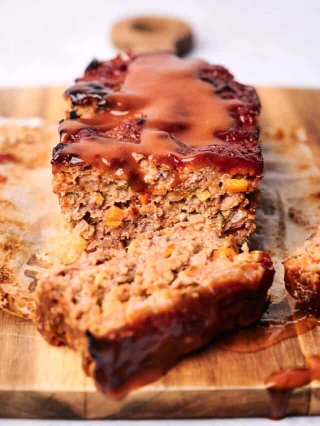 A meatloaf topped with a brown sauce, partially sliced, showing the interior with visible bits of vegetables, placed on a wooden cutting board.