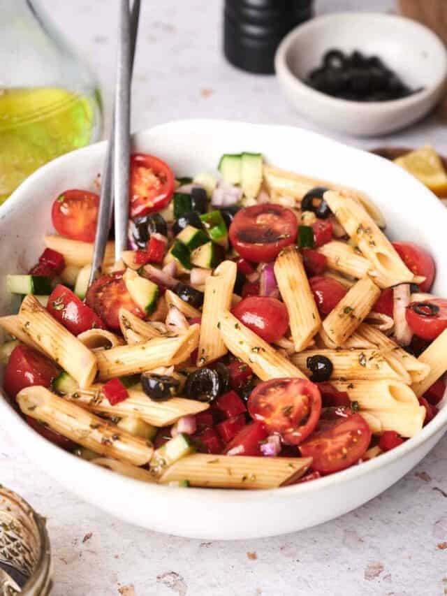 A white bowl filled with a pasta salad containing penne, cherry tomatoes, black olives, cucumber cubes, red onion, and herbs, with a spoon resting in it. A bottle of olive oil and condiments are in the background.