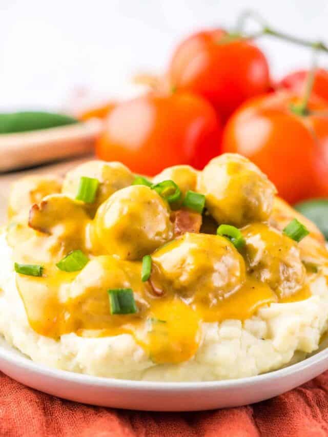 A plate of mashed potatoes topped with meatballs and a yellow sauce, garnished with chopped green onions. Fresh tomatoes are visible in the background.