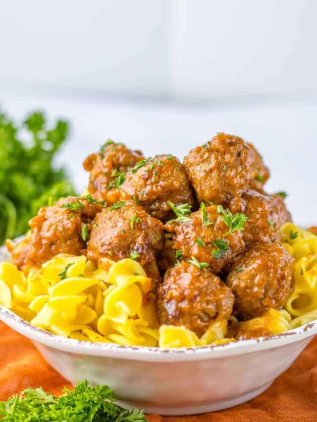 A bowl of swedish meatballs over egg noodles garnished with parsley, on a table with a vibrant orange cloth and greenery in the background.