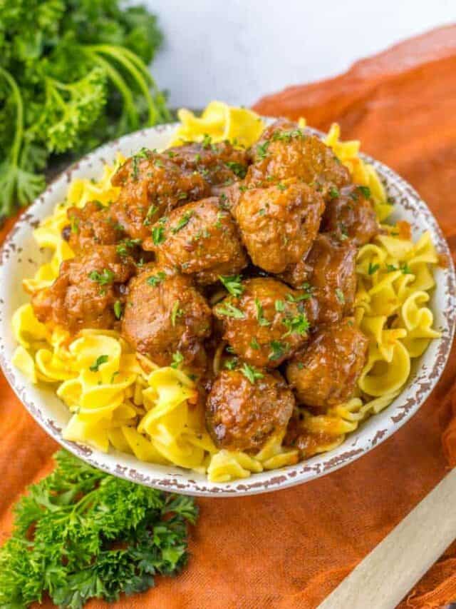 A bowl of egg noodles topped with saucy meatballs, garnished with chopped parsley, placed on an orange cloth with fresh parsley on the side.