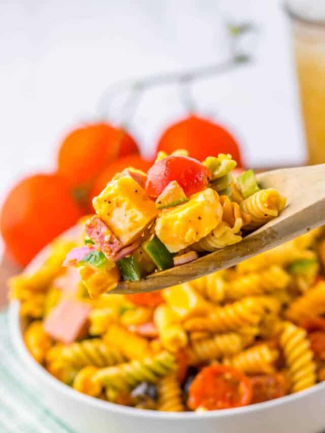 A spoonful of rotini pasta salad with diced vegetables, cherry tomatoes, and cheese is held above a bowl of the salad. Fresh tomatoes are seen in the background.
