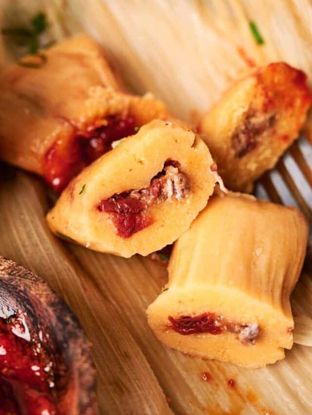 Close-up of sliced tamales filled with meat and red sauce, placed on a corn husk, with a wooden spoon partially visible in the lower left corner.
