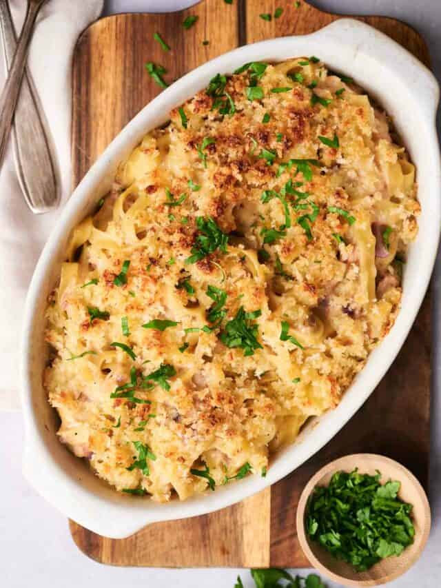 A baked noodle casserole topped with breadcrumbs and garnished with chopped parsley sits in a white dish on a wooden cutting board, with a small bowl of parsley and utensils nearby.