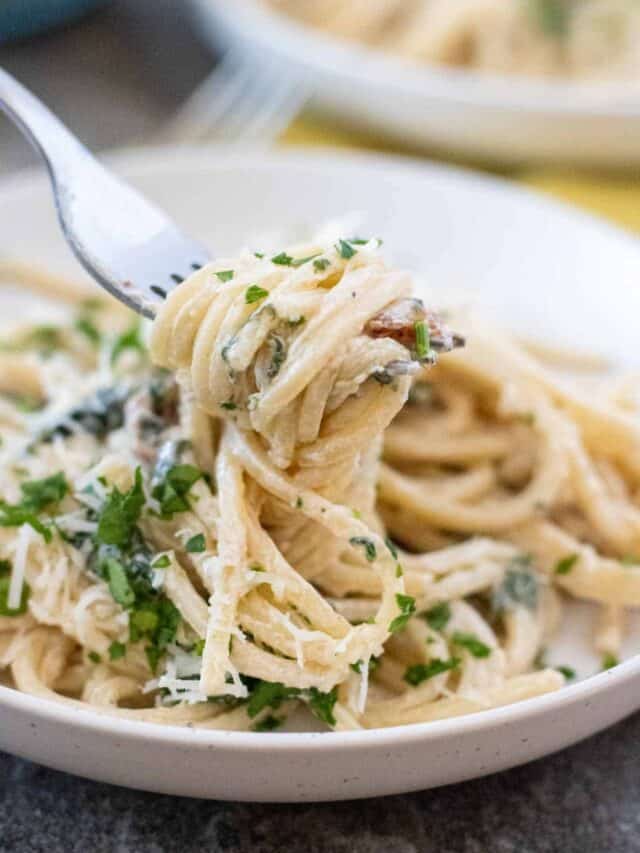 A fork lifts a portion of creamy spaghetti garnished with chopped parsley from a white plate.