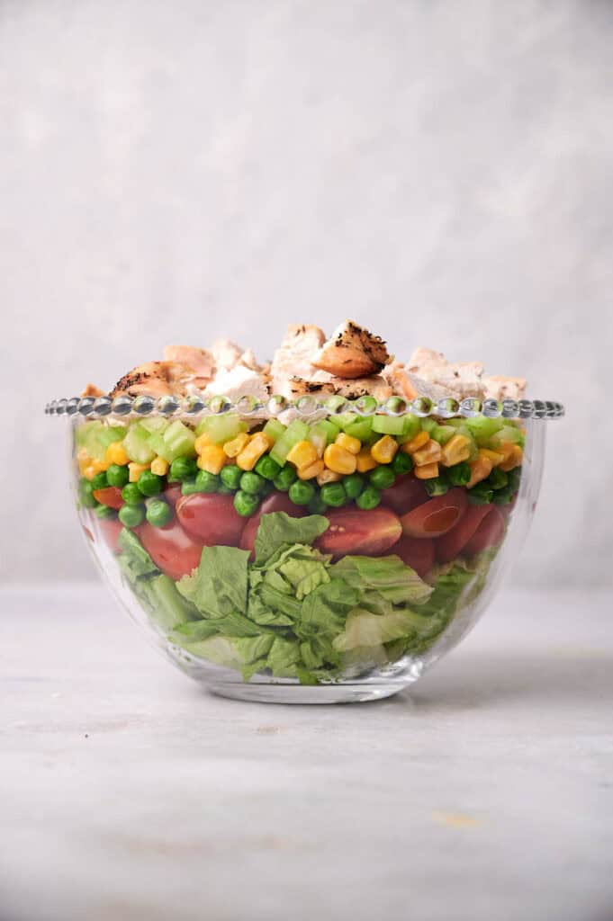 A clear glass bowl filled with layered salad including lettuce, tomatoes, peas, corn, and topped with grilled chicken pieces.