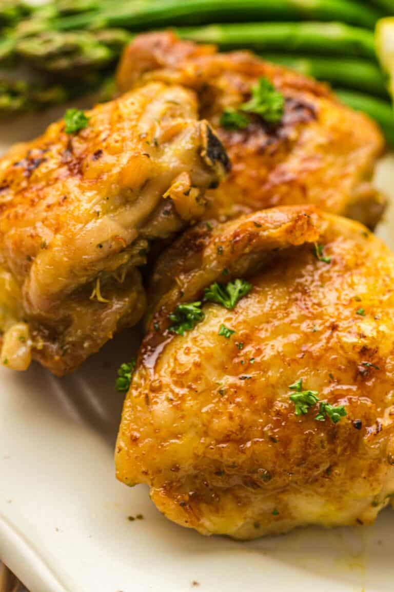 Close-up of two pieces of AIR FRIED chicken thighs garnished with parsley on a plate, with a side of asparagus in the background.