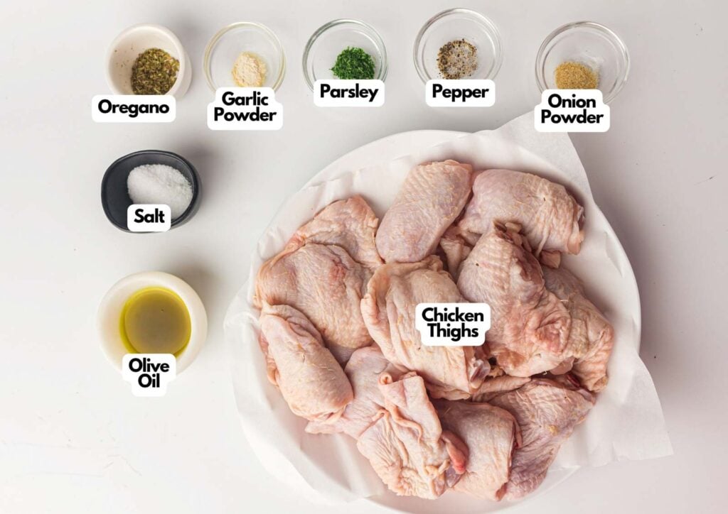A plate of raw chicken thighs is surrounded by small bowls containing oregano, garlic powder, parsley, pepper, onion powder, salt, and olive oil.