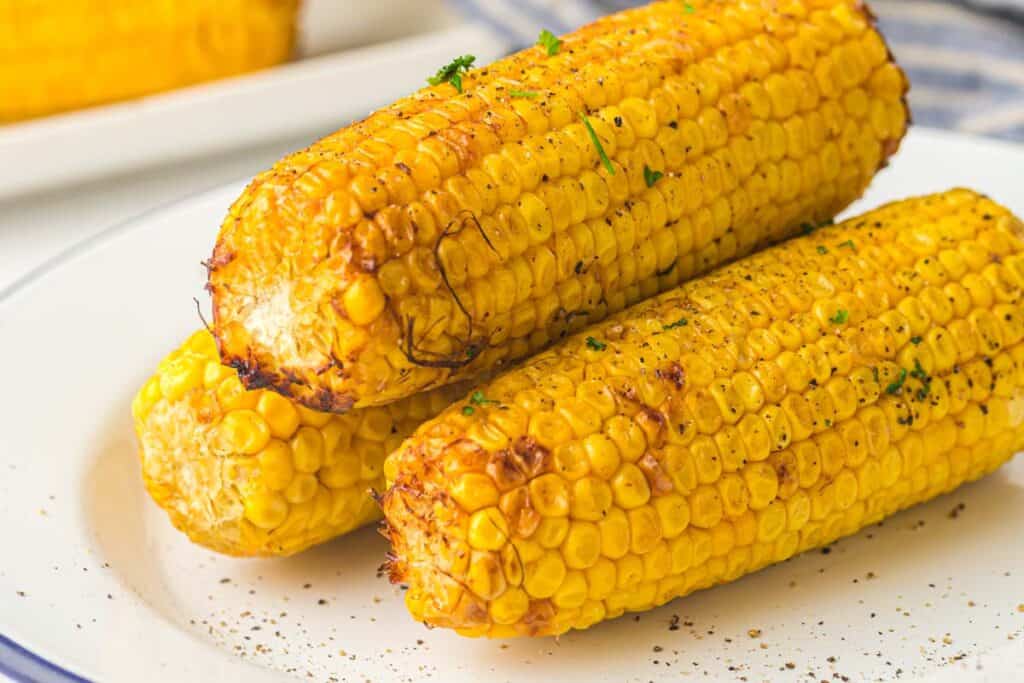 Three grilled corn on the cobs seasoned with black pepper and garnished with herbs are placed on a white plate.