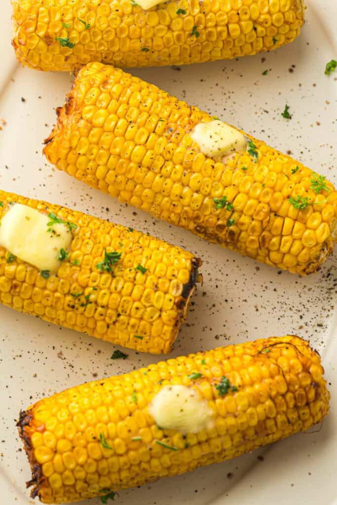 Four grilled corn on the cob pieces topped with melting butter and sprinkled with herbs on a white plate.