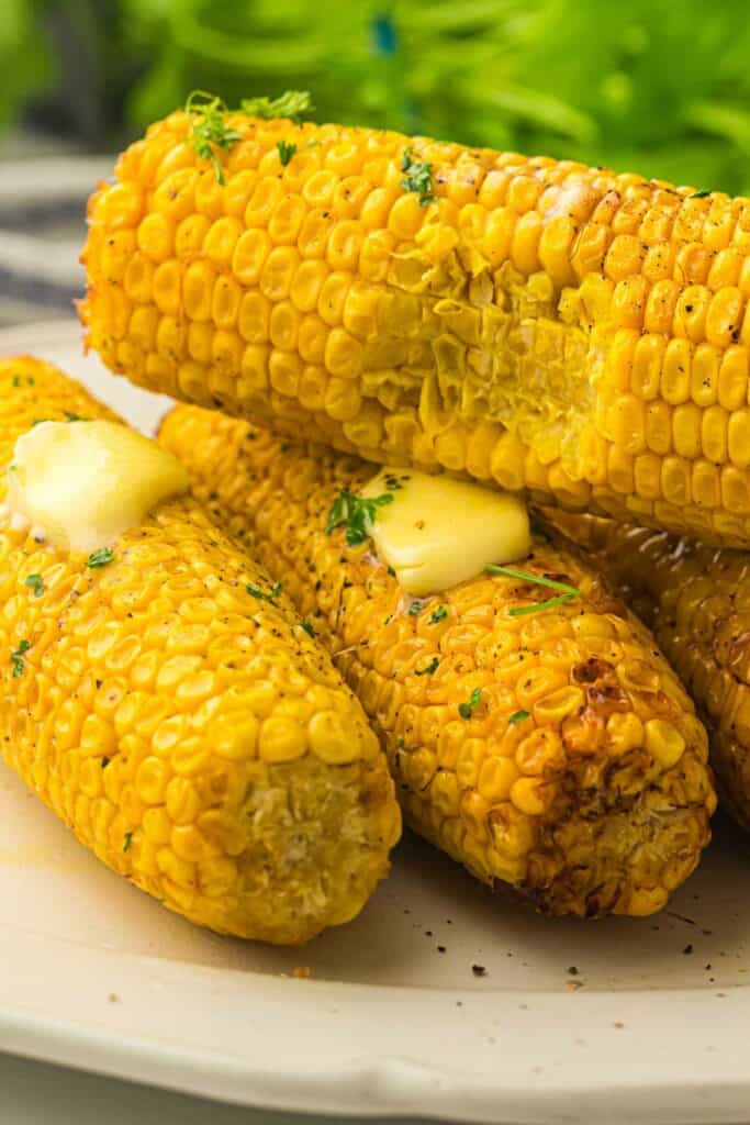 Three grilled corn on the cobs garnished with herbs and topped with melting pats of butter are placed on a plate.