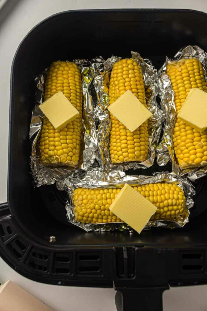 Four ears of corn with butter on top, placed in foil and arranged in a black air fryer basket.