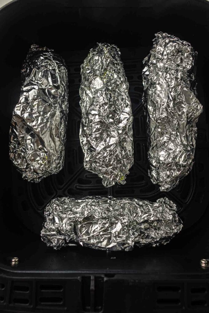 Four pieces of food wrapped in aluminum foil placed inside an air fryer basket.