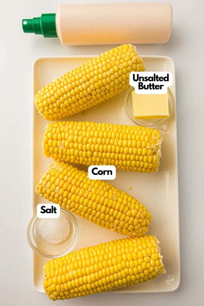 A rectangular plate holds three ears of corn next to small dishes of unsalted butter and salt, with a bottle of sauce in the background.