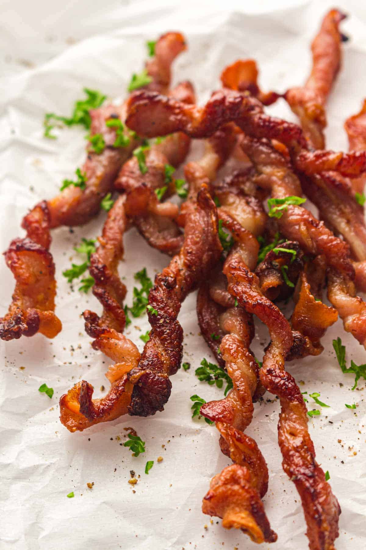 Crispy twisted bacon strips garnished with chopped parsley on parchment paper.