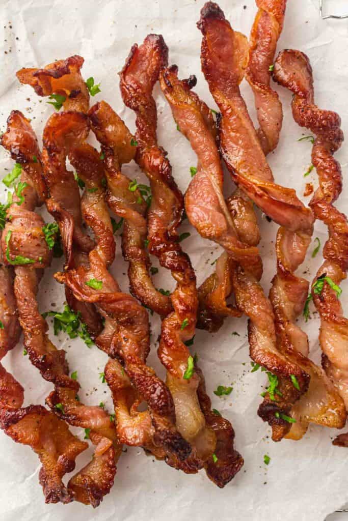 Crispy, twisted bacon strips garnished with chopped parsley on a white parchment paper background.