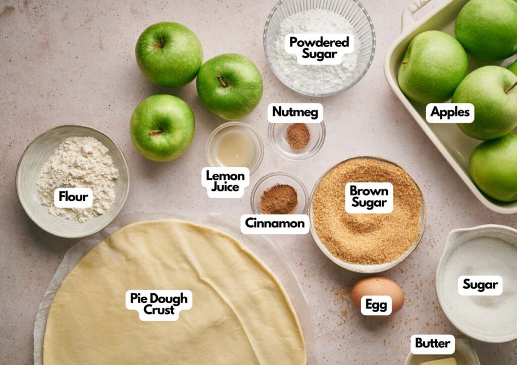 Labeled ingredients for classic apple pie, including green apples, flour, pie dough crust, lemon juice, cinnamon, nutmeg, brown sugar, egg, butter, sugar, and powdered sugar arranged on a countertop.