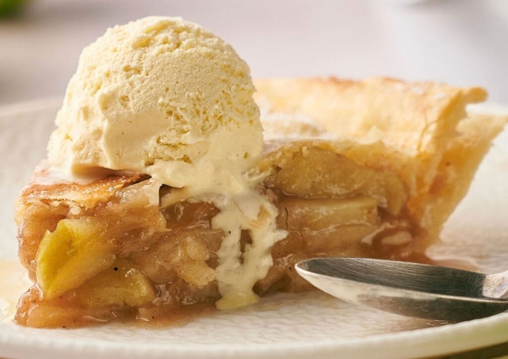 A slice of classic apple pie topped with a scoop of vanilla ice cream sits on a white plate next to a spoon.