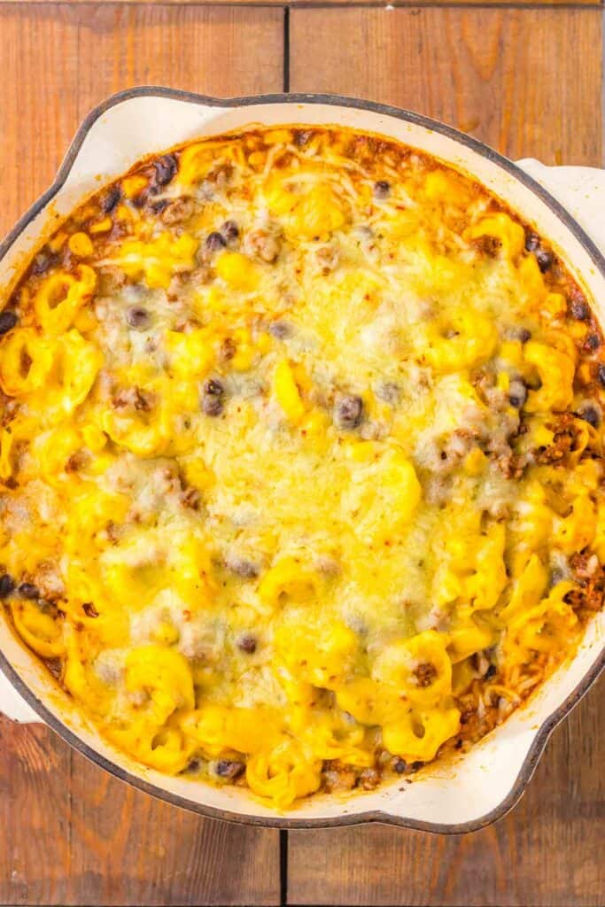 A baked dish filled with beef enchilada tortellini, melted cheese, and browned toppings in a round white pan on a wooden surface.