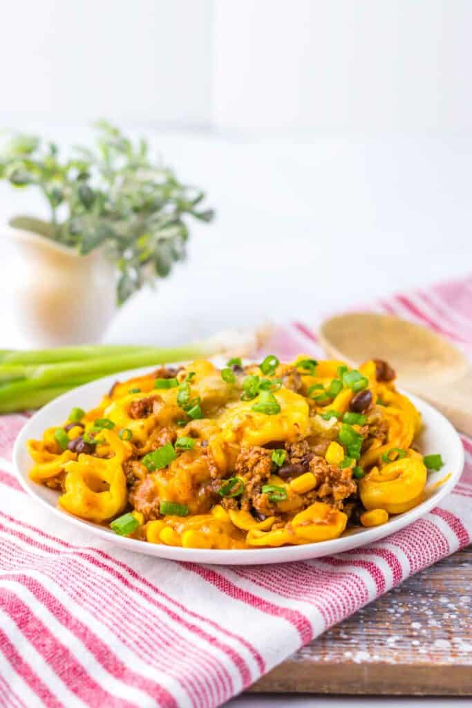 A plate of Beef Enchilada Tortellini topped with ground meat, black beans, corn, and chopped green onions, set on a red and white striped cloth with a wooden spoon and small potted plant in the background.