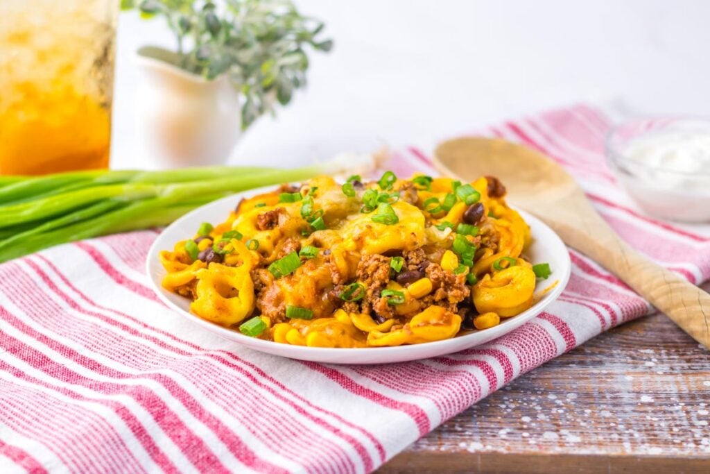 A plate of Beef Enchilada Tortellini features cheesy pasta with ground meat, black beans, corn, and green onions on a striped cloth, with a wooden spoon and fresh green onions nearby.