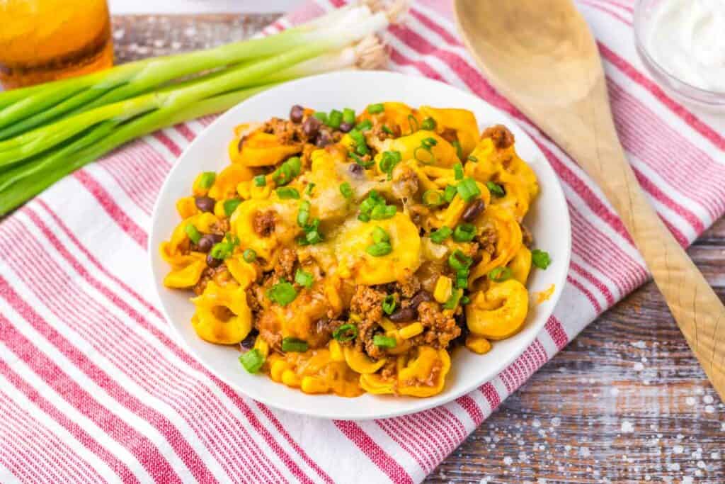 A plate of Beef Enchilada Tortellini topped with ground meat, cheese, black beans, corn, and green onions sits on a red and white striped cloth next to wooden spoons. Green onions and a glass jar are in the background.