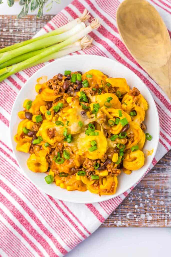 A plate of Beef Enchilada Tortellini mixed with cheesy tortellini pasta, ground meat, black beans, corn, and green onions, served on a red and white striped cloth next to several green onions and a wooden spoon.
