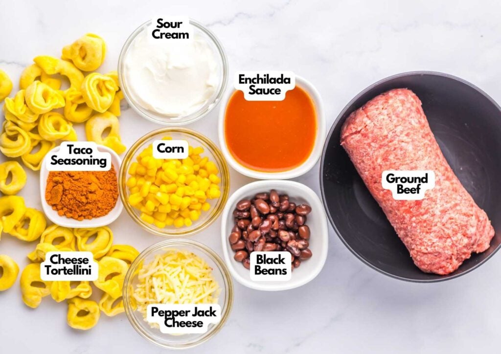 Ingredients for Beef Enchilada Tortellini on a white surface: cheese tortellini, taco seasoning, corn, pepper jack cheese, sour cream, enchilada sauce, black beans, and ground beef.