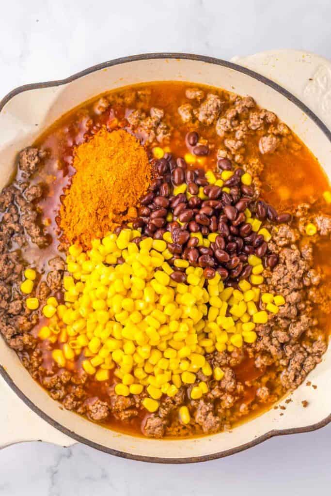 A skillet contains cooked ground beef, yellow corn, black beans, and a heap of spices, prepared for mixing into a Beef Enchilada-inspired dish.