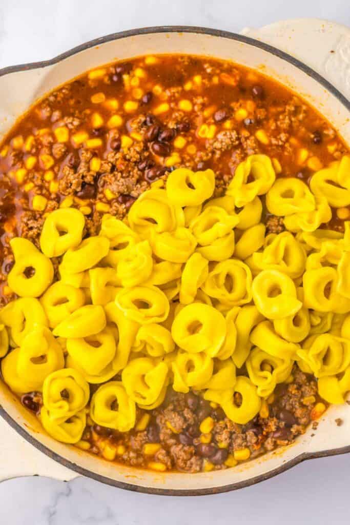 A skillet holds a savory blend of ground meat, black beans, corn, and tomato sauce on one side, reminiscent of beef enchilada flavors, while uncooked yellow tortellini pasta awaits its turn on the other side.
