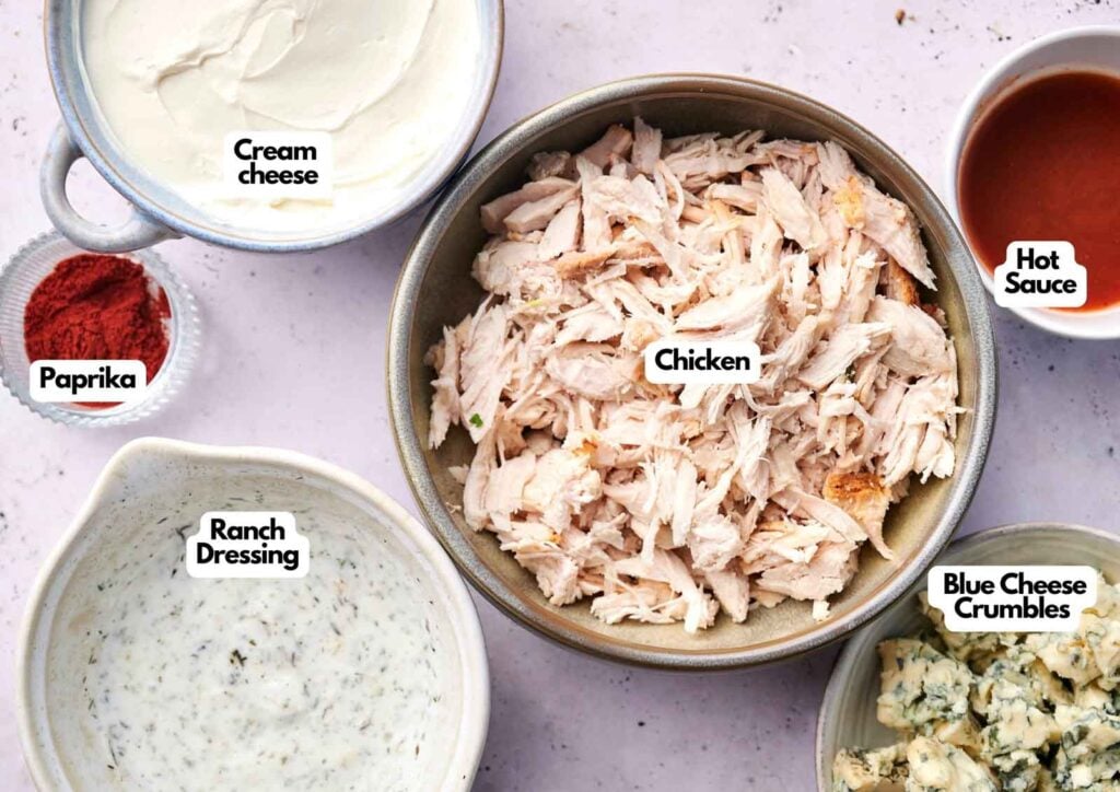 Bowls containing Buffalo Chicken dip ingredients like shredded chicken, cream cheese, ranch dressing, blue cheese crumbles, paprika, and hot sauce are arranged on a light surface, each ingredient labeled.