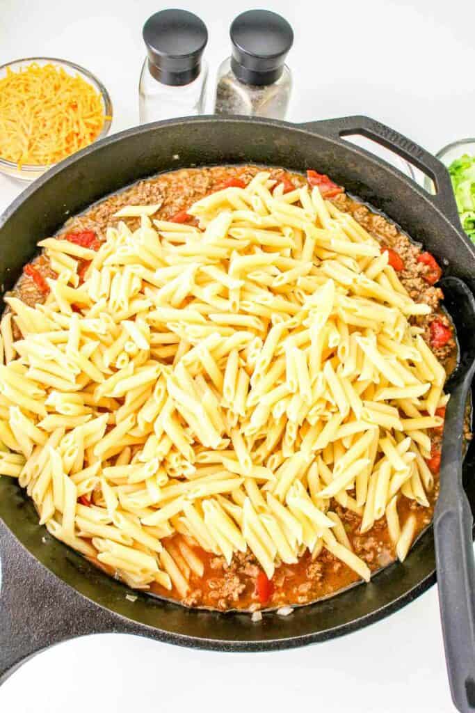 A skillet filled with cooked ground meat, tomatoes, and uncooked penne pasta, reminiscent of a cheeseburger pasta, with cheese and condiments in the background.