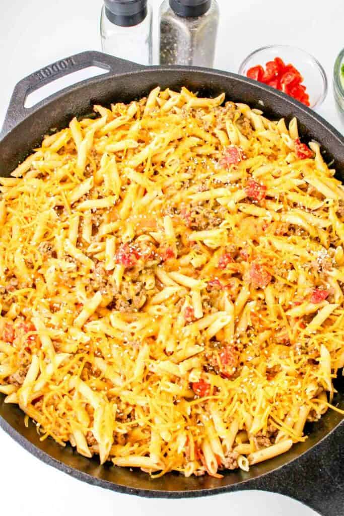 A skillet containing penne pasta mixed with ground meat and diced tomatoes, all topped with melted cheese for a delightful cheeseburger pasta twist.