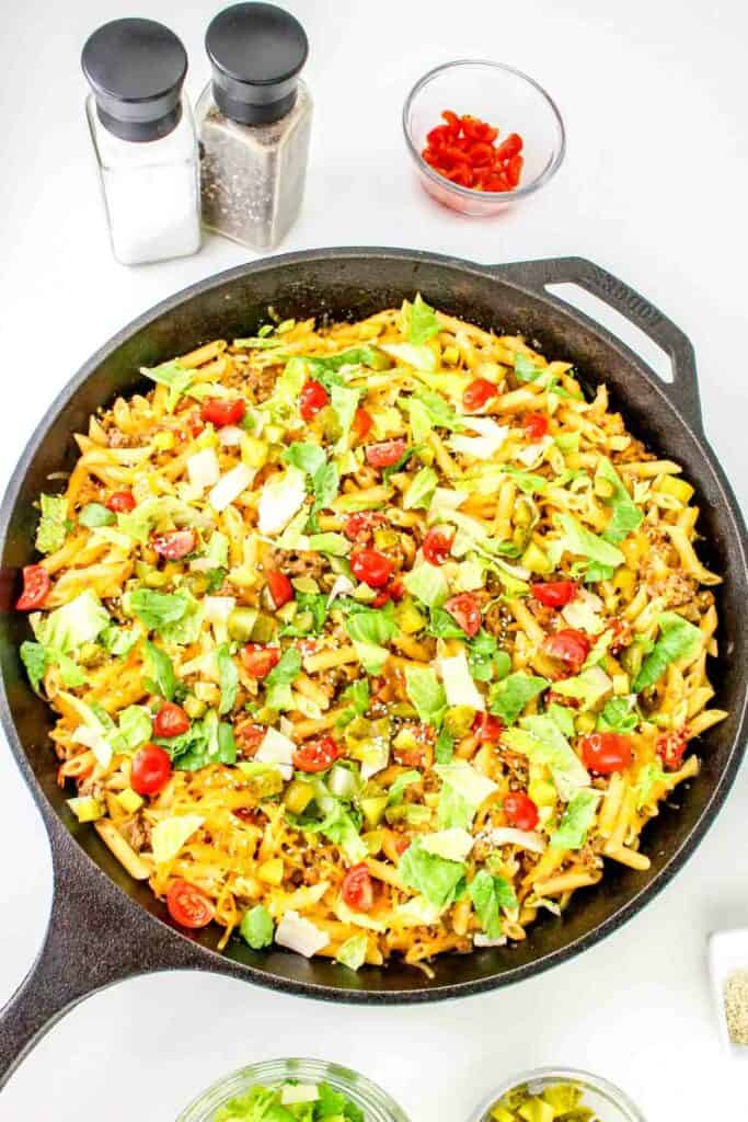 A skillet filled with cheeseburger pasta: cooked pasta mixed with ground meat, fresh cherry tomatoes, shredded cheese, and garnished with chopped greens. Two containers of salt and pepper, a bowl of sliced peppers, and a bowl of cherry tomatoes are in the background.